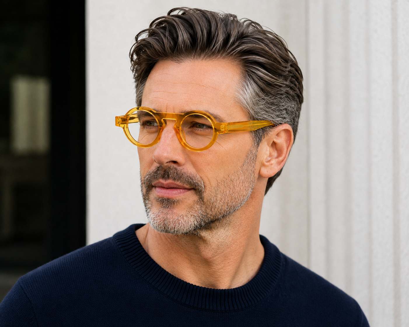 A man with styled gray hair and a beard wearing round amber-yellow translucent eyeglasses and a dark navy sweater, shown in a close-up portrait against a light architectural background.